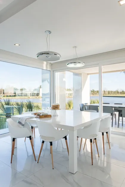 Comedor moderno blanco con vista al lago y sillas de madera — proyecto TRV 490, por Estudio Arq Daniel Tarrio