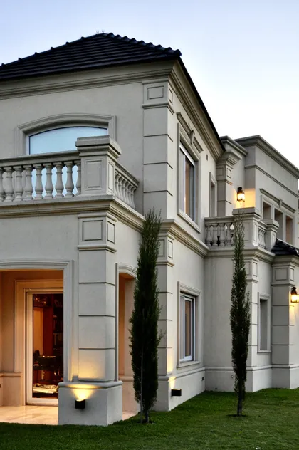 Casona clasica blanca con balcon, puerta naranja y jardin iluminado — proyecto Ayres del Pilar, por Marconi Silva Arquitectos, en Pilar