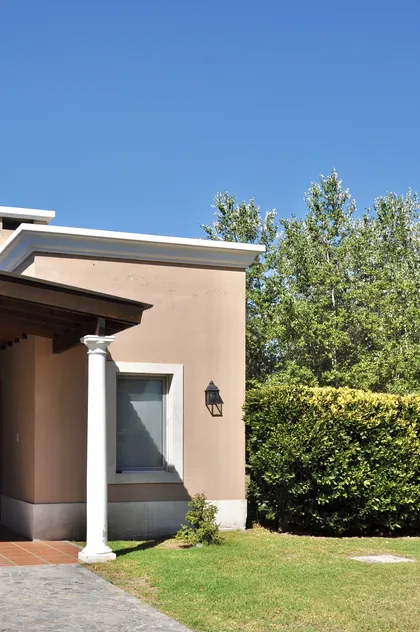 Fachada moderna terracota con columna blanca y jardín verde — proyecto Chacras de la Cruz, por Marconi Silva Arquitectos, en Capilla del Señor