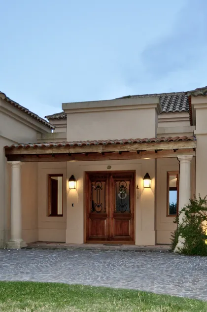 Fachada principal con portico de columnas, puerta de madera y piso empedrado — proyecto El Solar de Capilla, por Marconi Silva Arquitectos, en Capilla del Señor
