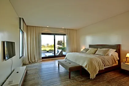 Dormitorio luminoso con ventanal, cama de madera y piso de parquet — proyecto Golf Club Nordelta, por Marconi Silva Arquitectos, en Nordelta