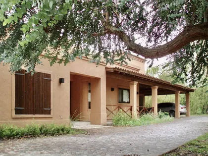 Casa de campo con fachada terracota, porche cubierto y frondoso árbol — proyecto La Retama, por Marconi Silva Arquitectos, en Manzanares