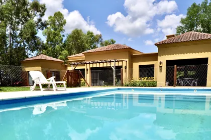 Casa amarilla con pileta celeste, deck y pergola, rodeada de árboles — proyecto La Retama 2, por Marconi Silva Arquitectos, en Manzanares