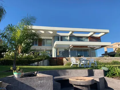 Fachada moderna de concreto blanco con ventanales en entorno costero — proyecto Casa Cardales, por Barrionuevo Arquitects, en Los Cardales
