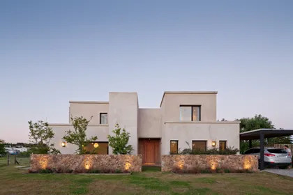Casa contemporánea blanca con fachada escalonada y jardín iluminado — proyecto La Tahona, por BAS Y GARCIA , en Canelones Departamento de Canelones