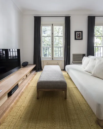Dormitorio moderno con cama blanca, banco gris y ventanal con cortinas negras — proyecto Arenales, por BAS Y GARCIA , en Recoleta