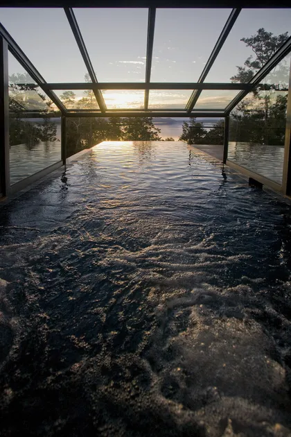 Piscina infinita interior con vista al atardecer y techo de cristal — proyecto Casa Muelle de Piedra, por Sidoni & Asociados, en Villa La Angostura