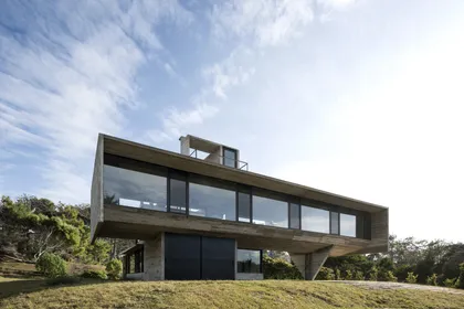 Casa moderna hormigón con grandes ventanales sobre terreno elevado — proyecto Casa Cariló, por Luciano Kruk, en Cariló