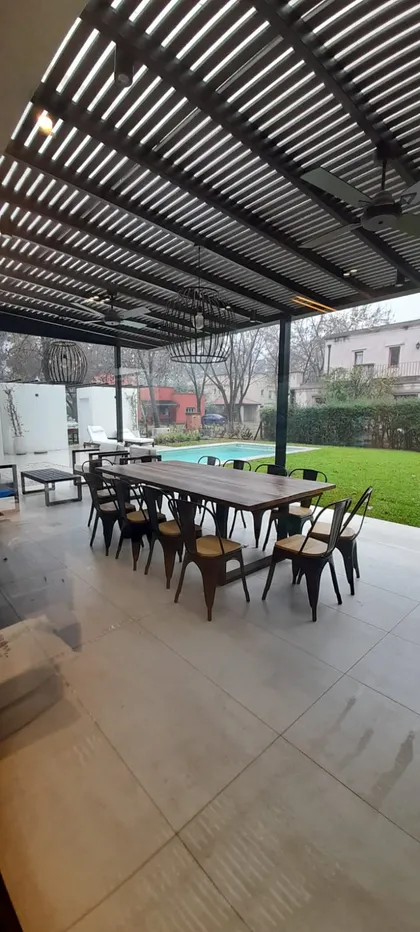 Parrilla cubierta moderna con mesa de madera y sillas de metal, vista a la piscina — proyecto El 54, por Valeria Damiani Arquitecta, en Pilar