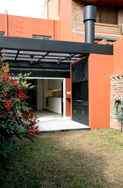 Quincho moderno con fachada naranja, pérgola negra y vegetación frondosa — proyecto Obra Martinez A, por Arq. Lourdes Yarade Saravia, en Martínez