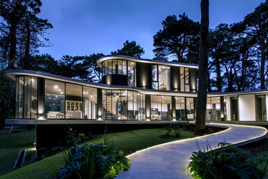 Casa moderna con fachada curva de vidrio en bosque nocturno — proyecto Casa Ameba, por MUCHTEK, en Cariló