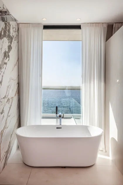 Baño moderno con bañera exenta frente a ventana con vista al mar — proyecto CASA MUELLES 239, por ABAD GADANO, en Belén de Escobar