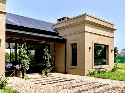 Fachada moderna beige con techo negro y grandes ventanales en jardín — proyecto Los Jazmines Pilar del Este, por Marconi Silva Arquitectos, en Pilar del Este