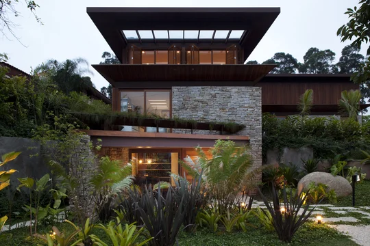 Casa contemporánea con fachada de piedra, grandes ventanales y jardín frondoso iluminado — proyecto Residência MN, por Gil Fialho