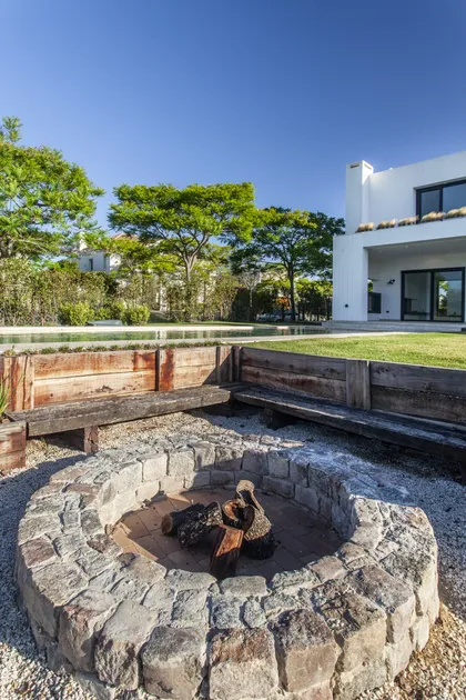Patio moderno con hogar de piedra, piscina y casa blanca al fondo — proyecto El Golf 137 - Nordelta, por Build a Home Arquitectura, en B1624 Tigre