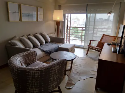 Living moderno con sofá seccional, sillón y ventanales amplios — proyecto Departamento RA, por Vicky Hurrell, en Mar del Plata