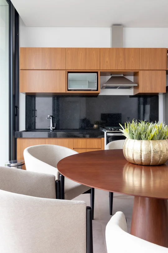 Comedor con mesa de madera redonda y cocina integrada con muebles de madera — proyecto Uso Misto Anza, por Studio Jordano Valota