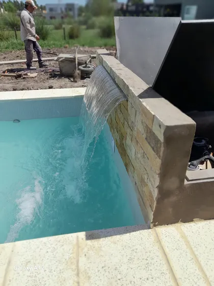 Pileta de obra en construcción con agua turquesa y cascada — proyecto piscina con playa humeda mas cascada, por Piscinas omar, en Canning