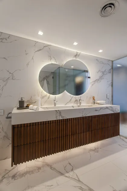 Baño moderno con doble lavabo, espejo curvo retroiluminado y mueble de madera — proyecto Pacífico BRT, por Vanguarda Arquitectos, en CABA