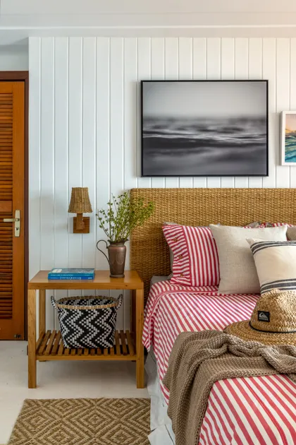 Dormitorio luminoso con pared blanca en tablillas, cama de ratán y ropa de cama rayada rojo blanco — proyecto Casa da Ilha, por Miguel Capanema Arquitetura