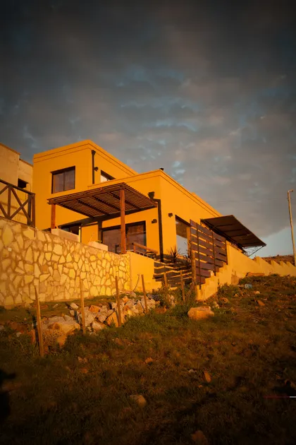 Casa moderna amarilla con pérgola y muro de piedra en ladera — proyecto Inmuebles, por Geo Estudio, en Buenos Aires