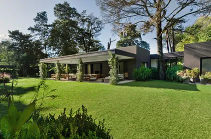 Moderna fachada de casa con jardín exuberante y grandes ventanales — proyecto Casa M, por Estudio Dado, en Pilar