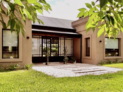 Fachada moderna con ventanales negros, patio de grava blanca y jardín frondoso — proyecto Los Jazmines Pilar del Este, por Marconi Silva Arquitectos, en Pilar del Este