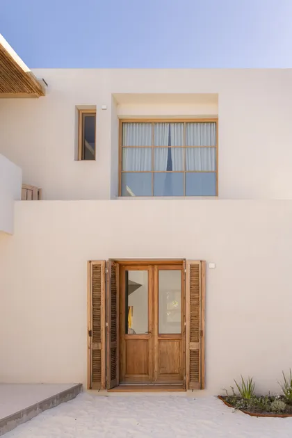 Fachada blanca minimalista con puertas de madera y persianas — proyecto El Refugio, por Grupo Pauta Arquitectura, en Pinamar