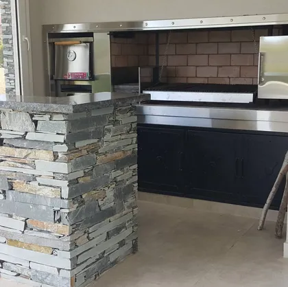 Pilar de piedra apilada y cocina exterior moderna con parrilla integrada — proyecto LA RESERVA CARDALES - LRCC 195, por LESSI ARQUITECTURA, en Los Cardales