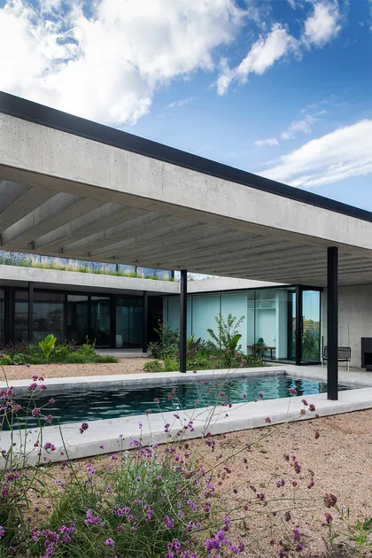 Casa moderna con pileta bajo pérgola, flores moradas y cielo despejado — proyecto CASA CONSERVATORIO, por GMARQ, en Belén de Escobar