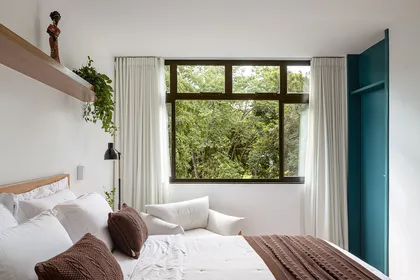 Dormitorio luminoso con ventanal hacia jardín, cama blanca y puerta verde — proyecto Apartamento Cobogó, por Coda Arquitetos