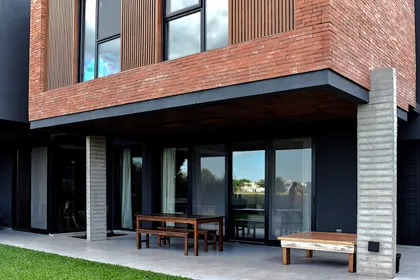 fachada moderna de ladrillo con grandes ventanales y mobiliario exterior de madera — proyecto CASA HC, por Daniel Daniele - Oficina de Arquitectura, en Canning