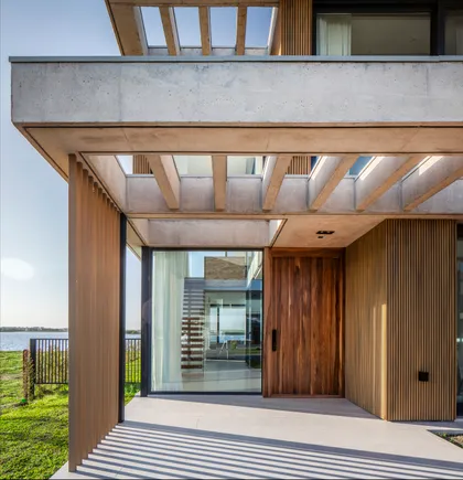 Fachada moderna con puerta pivotante de madera y detalles de hormigón — proyecto CASA MUELLES 239, por ABAD GADANO, en Belén de Escobar