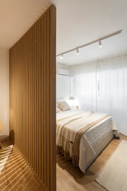 Dormitorio moderno con divisor de madera, cama de lino blanco y luz cálida — proyecto REFORMA NE, por Rastro Estudio, en Palermo