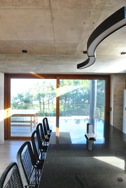 Cocina moderna integrada con isla, techos de hormigón y amplios ventanales — proyecto Casa FSC, por TAVA Coop, en Pinamar