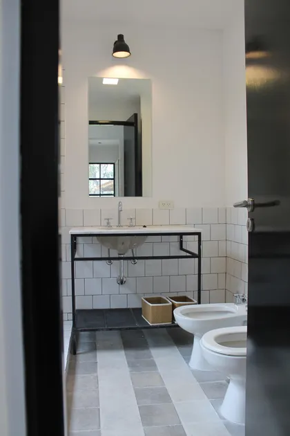 Baño industrial con bacha de pedestal, azulejos blancos y estanterías negras — proyecto Casa Recodo, por Ronda Arquitectura, en Garin