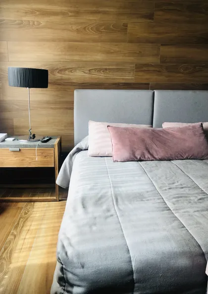 Dormitorio moderno con pared de madera, cama gris y luz cálida — proyecto SANTA BARBARA , por Greschek Obras , en Benavidez