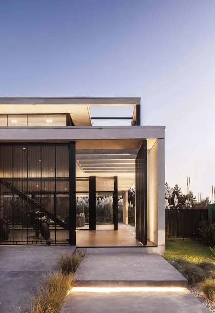 Fachada moderna de concreto con iluminación indirecta y grandes ventanales — proyecto Casa Barbarita, por Ezequiel Amado Cattaneo - EAC, en Troncos del Talar