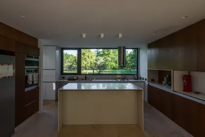 Cocina moderna con isla y vista al bosque a través de amplios ventanales — proyecto CASA AC , por BUSCETTI & DI BERNARDO arquitectas, en Villa Elisa