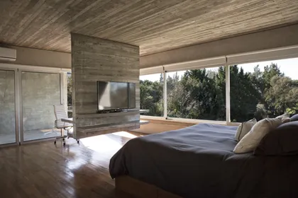 Dormitorio minimalista con cama negra, chimenea hormigón y vistas al bosque — proyecto Casa Torcuato, por Luciano Kruk