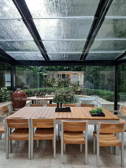 Comedor exterior moderno bajo techo de vidrio con vegetación abundante — proyecto TOLDER - CERRAMIENTOS, por Argentina en Obra 2024, en Rincón de Milberg
