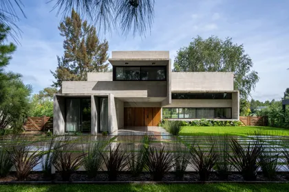 Fachada moderna de hormigón con grandes ventanales y jardín frondoso — proyecto Casa Miralagos II, por Besonías Almeida arquitectos, en La Plata