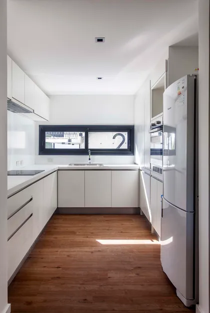 Cocina moderna estrecha con gabinetes blancos y piso de madera oscura — proyecto Alvear AV, por V2 Arquitectos, en City Bell
