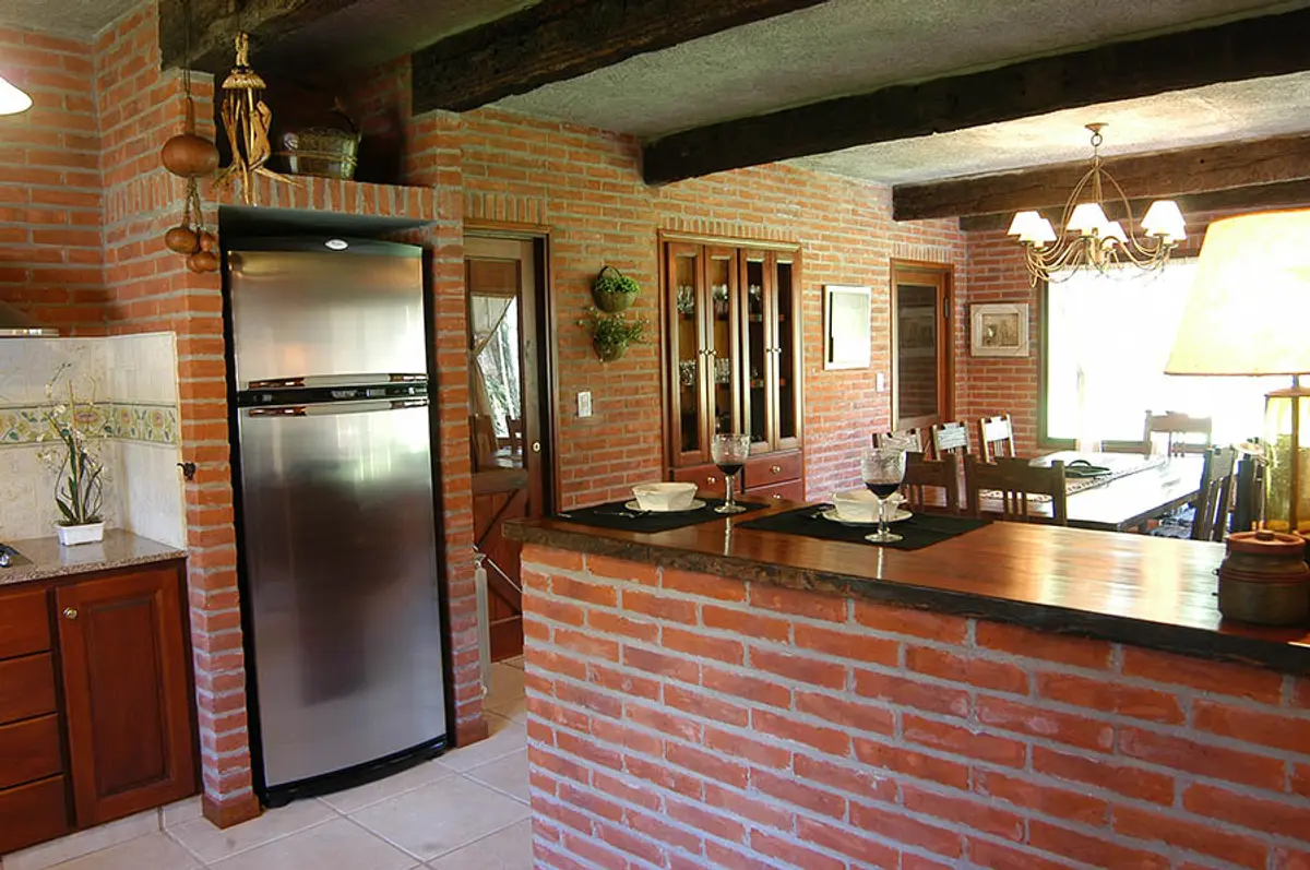 Cozinha rústica com paredes de tijolo aparente e vigas de madeira — projeto Casa Cariló 2004, por Hugo Zawadski Arquitectos, em Cariló