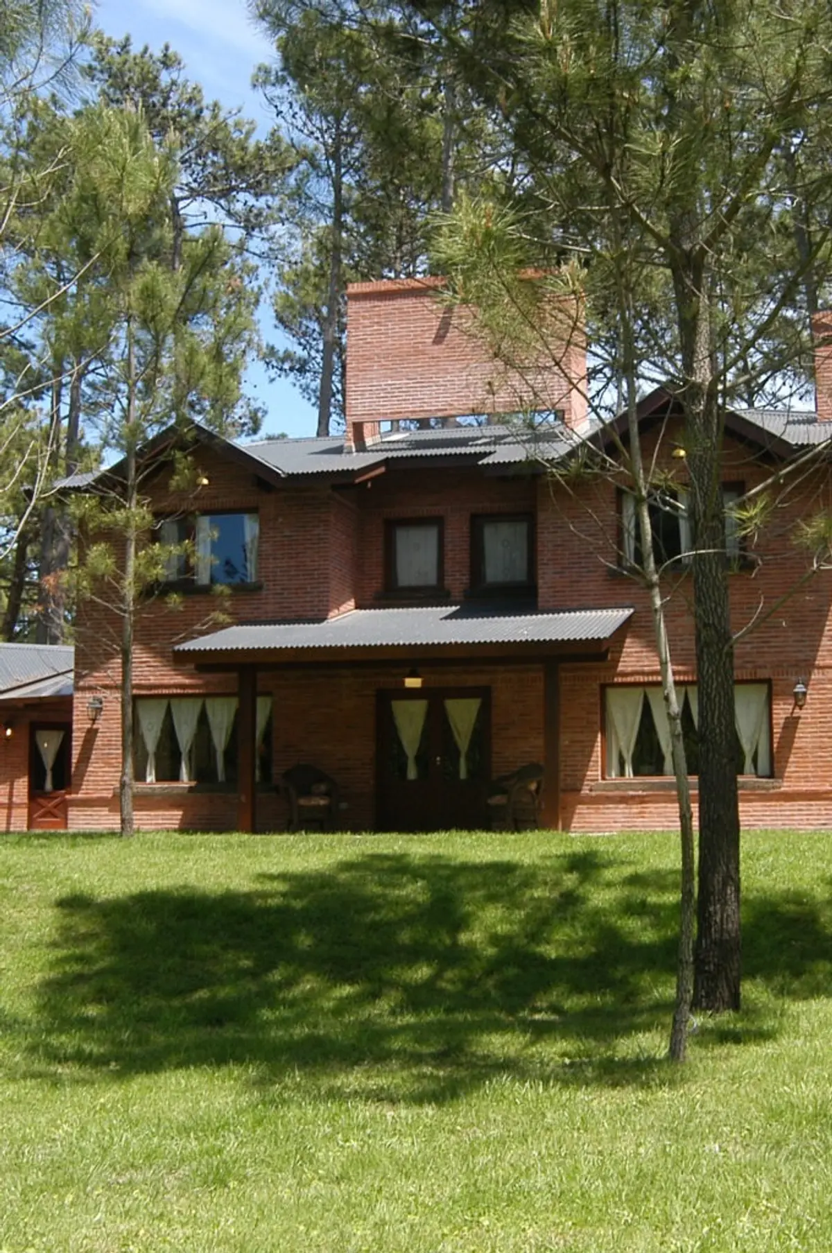 Fachada rústica de tijolos com pinheiros e gramado — projeto Casa Cariló 2004, por Hugo Zawadski Arquitectos, em Cariló