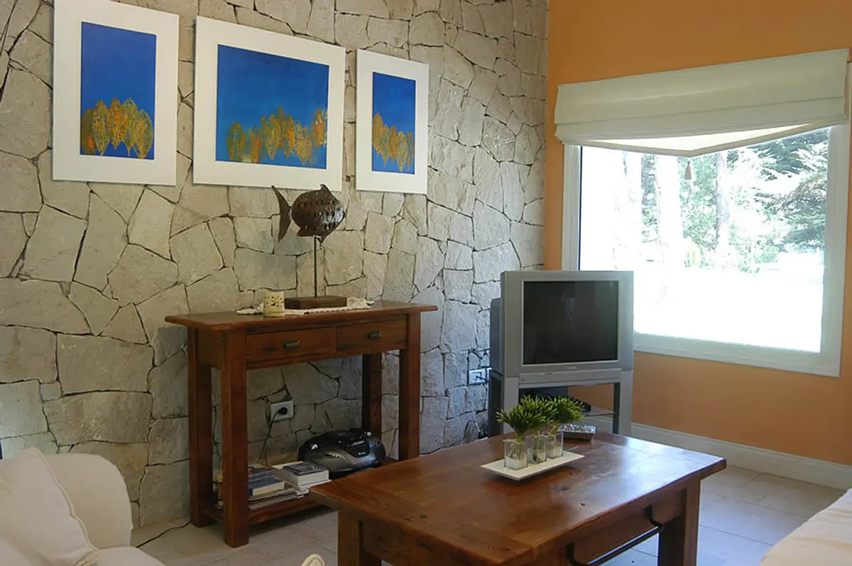 Sala de estar e jantar com parede de pedra e móveis de madeira — projeto Casa Cariló 2005, por Hugo Zawadski Arquitectos, em Cariló
