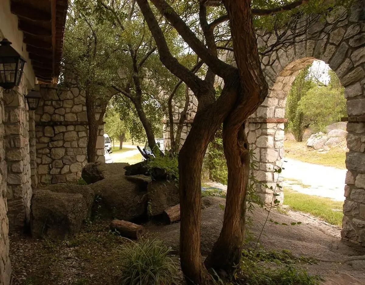 Pátio rústico com arcos e muros de pedra cercado por vegetação exuberante — projeto Paisajismo Aire, por Ricardo Pereyra Iraola