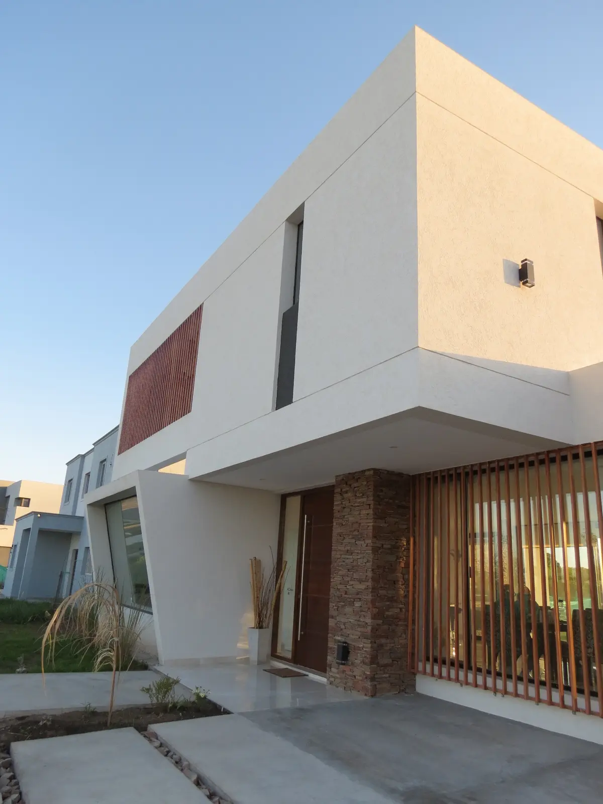 Fachada moderna con revestimiento blanco, pantalla de madera y detalles de ladrillo — proyecto Casa MB- Harás Sta. María , por Olivieri Arquitectura, en Haras Santa María