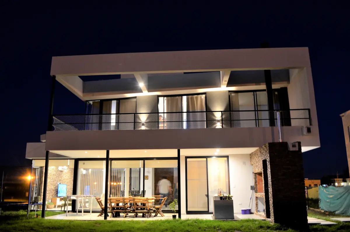 Casa moderna com fachada branca de concreto e grandes janelas, iluminada à noite — projeto Casa MB- Harás Sta. María, por Olivieri Arquitectura, em Haras Santa María