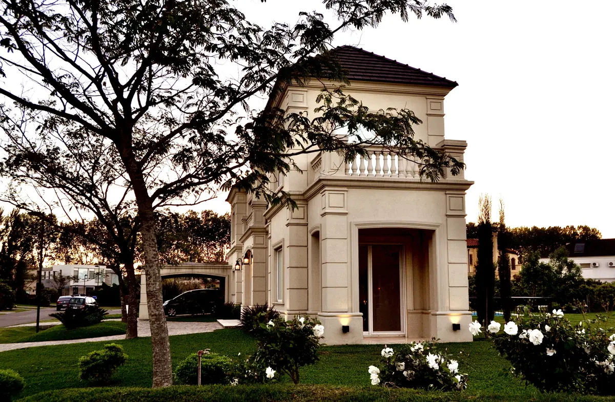 Fachada neoclásica blanca con jardín y árbol decorativo — proyecto Ayres del Pilar, por Marconi Silva Arquitectos, en Pilar
